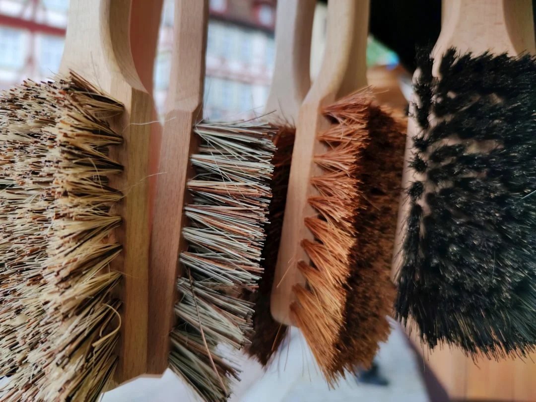 A flat lay of kolinsky sable watercolor rounds, hog-bristle oil brushes, and Japanese makeup brushes arranged on a wooden surface beside a watercolor palette and a Mason Pearson hairbrush